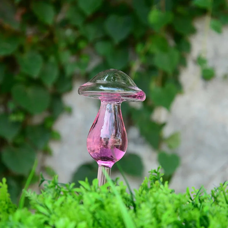 A clear, mushroom-shaped automatic drip watering device with a pink tint is inserted into a plant with lush green foliage, set against a backdrop of more greenery.