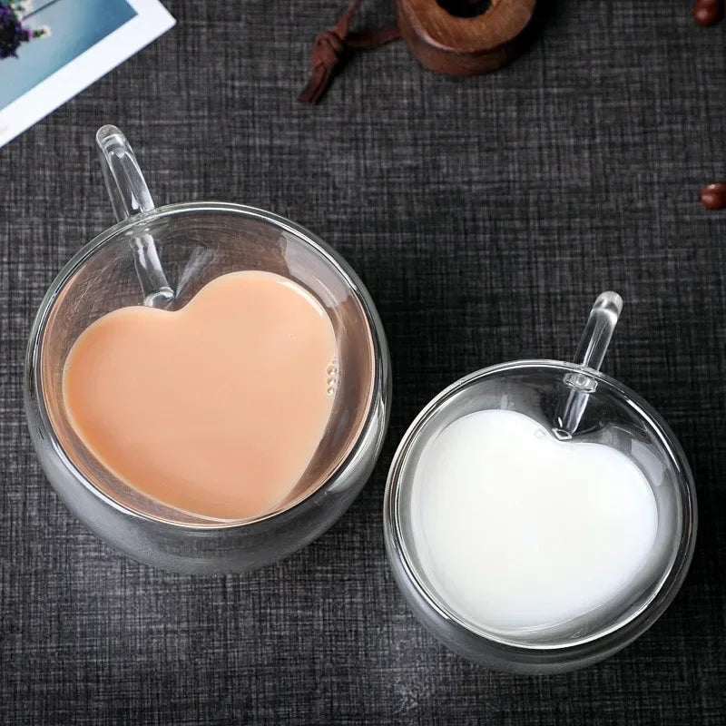 Two heart-shaped glass mugs are on a dark surface, one filled with coffee and the other with milk.