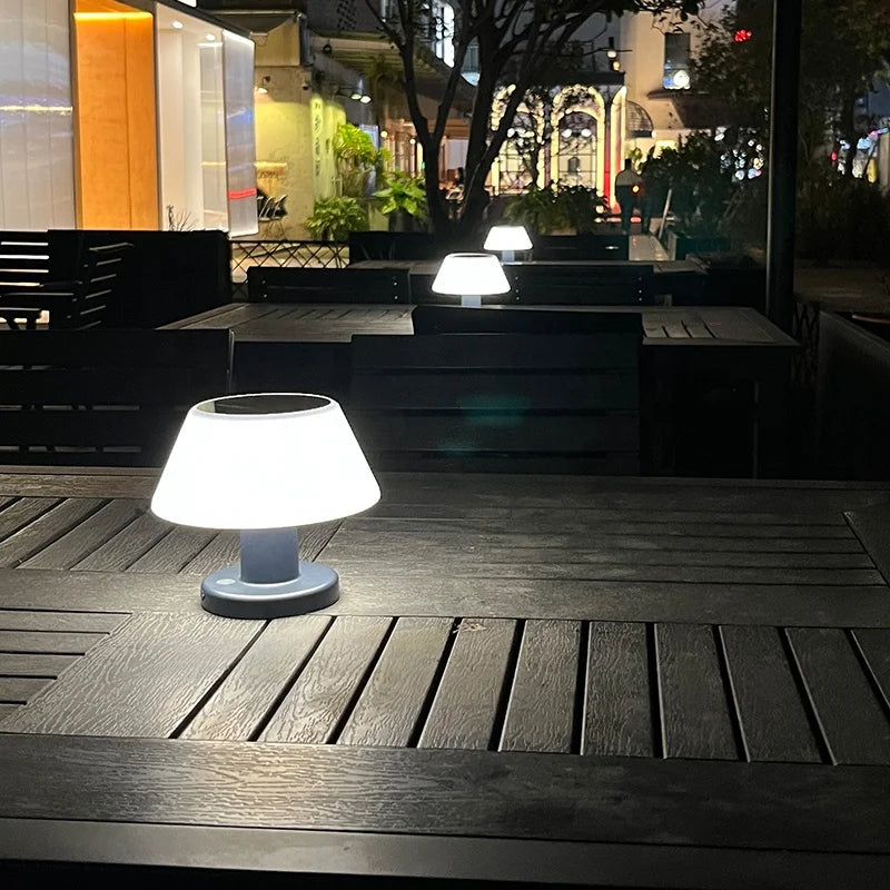 Three small, solar-powered table lamps with white, slightly tapered shades and dark bases illuminate dark wooden tables on an outdoor patio at night. The lamps cast a soft glow on the tabletops. The background shows blurred lights from nearby buildings and greenery.
