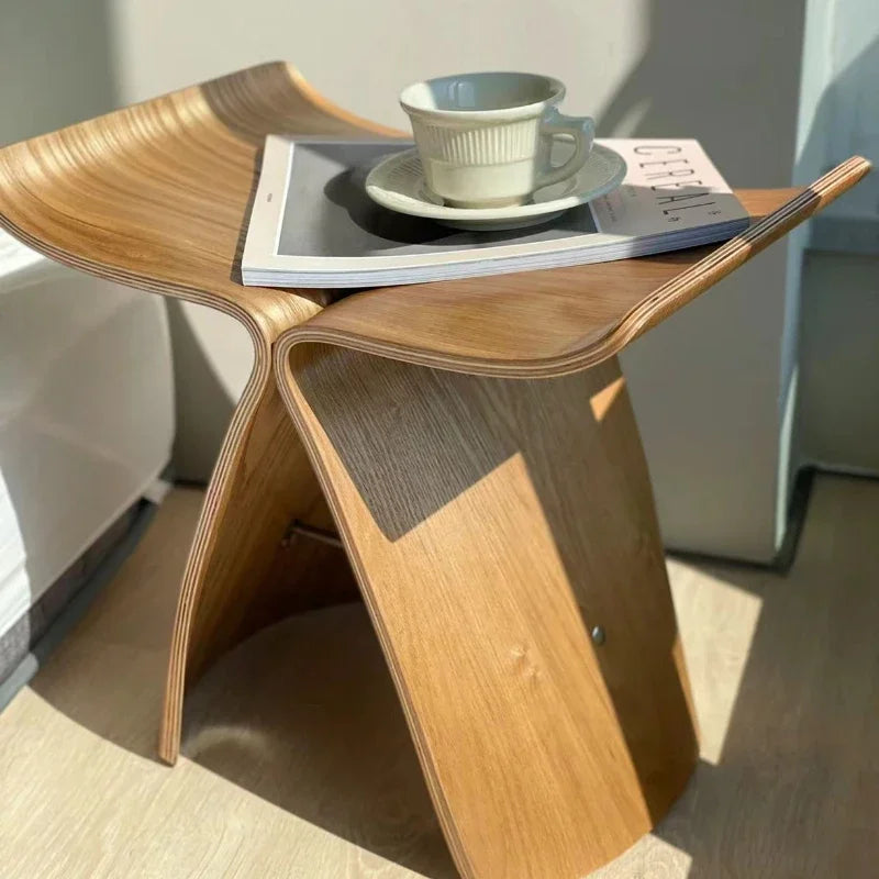 A light brown Hans Wegner Butterfly Stool, illuminated by sunlight, holds a white teacup and saucer on top of a magazine. The stool sits on a light-colored wooden floor. A portion of a light-colored sofa is visible in the blurred background.