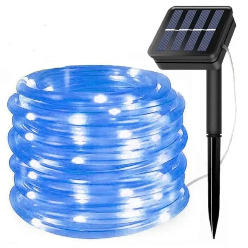 A coiled length of clear rope light emitting a bright blue glow is shown with its attached solar panel and ground stake against a white background. The rope light is neatly wound in a spiral.