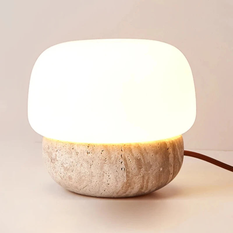 A small, glowing table lamp with a rounded white glass shade and a textured, light-colored stone base sits on a light surface. The lamp emits a soft, warm light, and its brown cord extends to the right. The background is a muted beige.