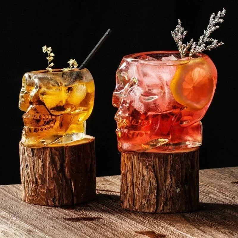 Two different colored skull-shaped glasses, one with a yellow-orange drink and the other with a red drink, both resting on wood log coasters.