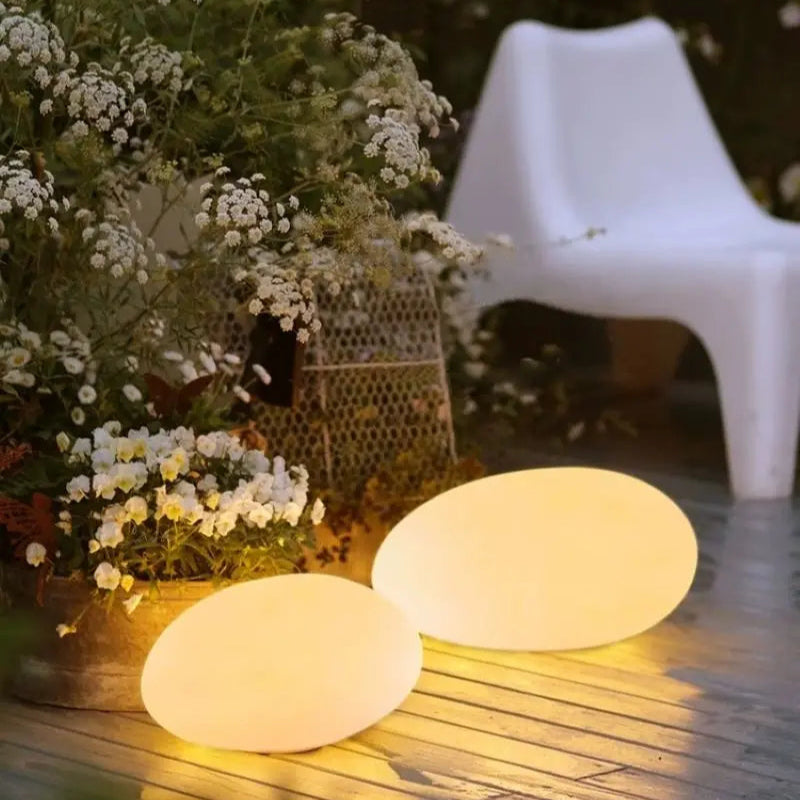 Two illuminated, round egg-shaped lights on a wooden patio, with a white modern chair and white flowering plants in the background.