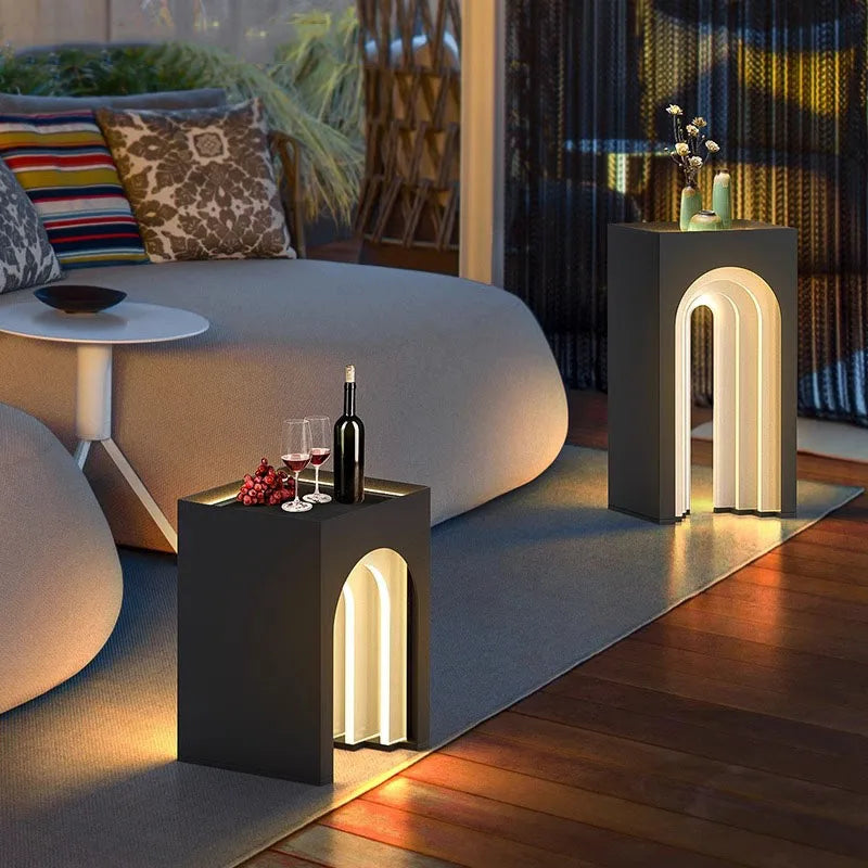 Two modern, dark gray solar arch lanterns illuminate an outdoor lounge area at twilight; one serves as a side table with wine and glasses.