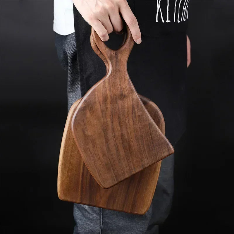 A person wearing a black apron and jeans holds two wooden cutting boards with circular handle cutouts. The boards are stacked, showcasing their different sizes and the natural wood grain. The person's hand grips the handle of the top board.