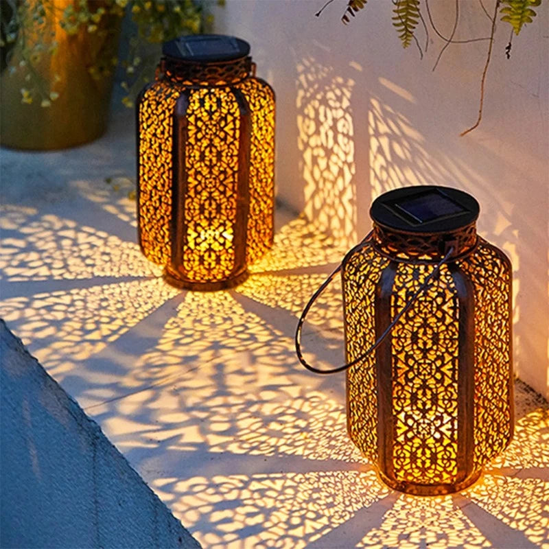 Two decorative metal solar lanterns with intricate cutouts emit a warm glow, casting patterned shadows on a light-colored surface at night. One lantern has a handle. Plants are visible in the blurred background.