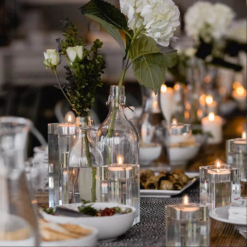 A festively set table features multiple clear glass candle holders, each with a lit tea light candle, interspersed with clear glass vases holding white flowers and greenery. A patterned runner lies on the table, which also holds white bowls of food, plates, and clear drinking glasses. The warm glow of the candles creates a cozy atmosphere.