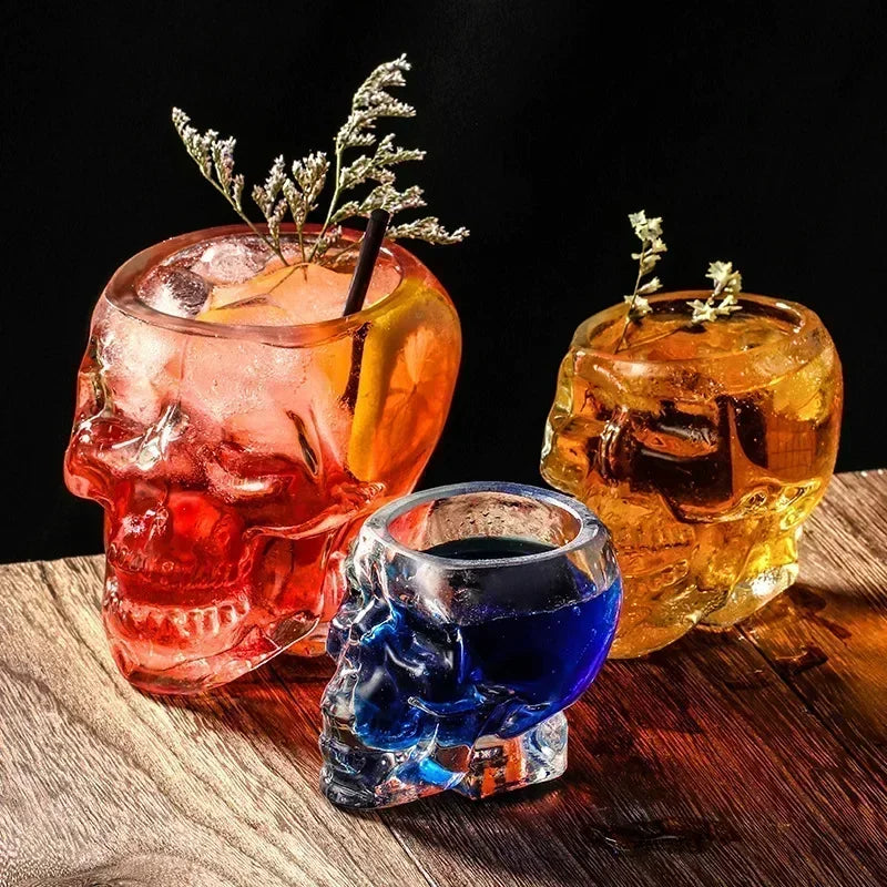 Three clear glass skull-shaped cups filled with different colored drinks, on a wooden surface. The drinks are a reddish-orange cocktail with a garnish, a small blue shot, and a large golden drink.