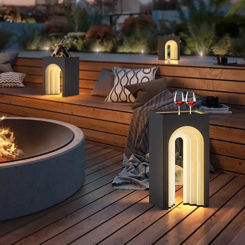 The image depicts a cozy outdoor lounge area at twilight. Two modern black side tables with integrated arched lighting illuminate the wooden deck. A round fire pit with flickering flames sits to the left. Comfortable seating with pillows and blankets invites relaxation, and two wine glasses rest on one of the illuminated tables. Soft outdoor lighting enhances the ambiance.
