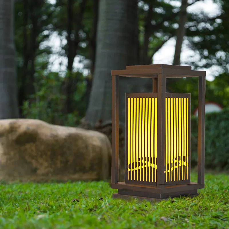 A modern, dark-framed outdoor lantern with vertical slats emits a warm yellow glow as it stands on green grass in a park-like setting. A large rock and several trees are visible in the blurred background.
