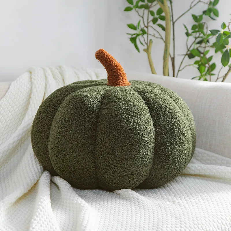 A dark green, fuzzy, pumpkin-shaped pillow with an orange-brown stem rests on a white textured blanket on a couch.