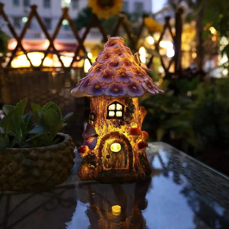 A small, whimsical fairy house with a textured brown base resembling a tree trunk and a conical roof adorned with purple and yellow flower-like details sits on a surface. The house features a small blue arched doorway with green moss accents and a blue window with a grid pattern. Miniature red and white spotted mushrooms grow along its sides.