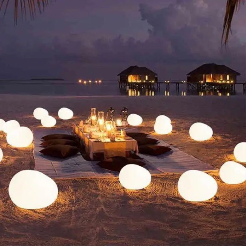 Romantic beach dinner setup at night with a table, pillows, and glowing egg-shaped lights on the sand, with bungalows in the distance.