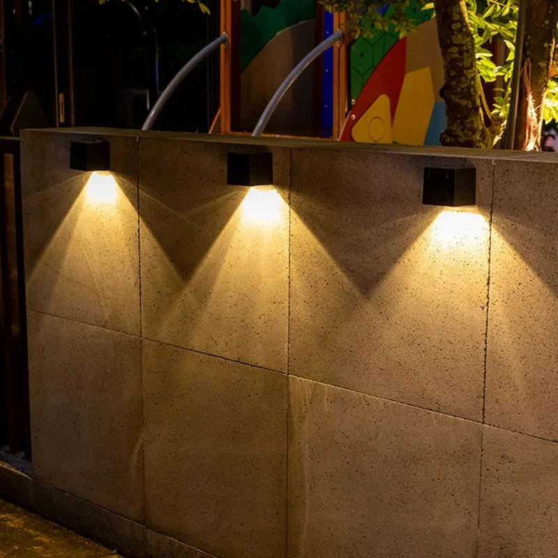 Three black solar-powered square wall lamps are mounted on a concrete wall, casting warm downward lighting in a triangular pattern. The outdoor setting includes a colorful background with trees and a playground structure.