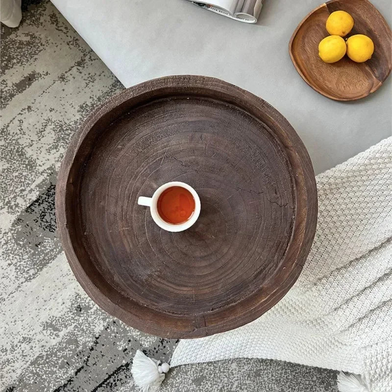 A top-down view of a round, dark wood, rustic side table with a shallow tray top, holding a small white cup of tea, with a partial view of a sofa, rug, and a plate of lemons.