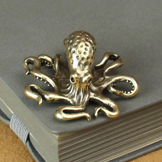 A small metallic octopus figurine with an antique brass or bronze finish, placed on top of a closed gray notebook. The octopus features intricate detailing, including textured tentacles and a sculpted head, giving it a vintage or steampunk aesthetic. The object appears to be decorative, possibly a charm, ornament, or small desk accessory.