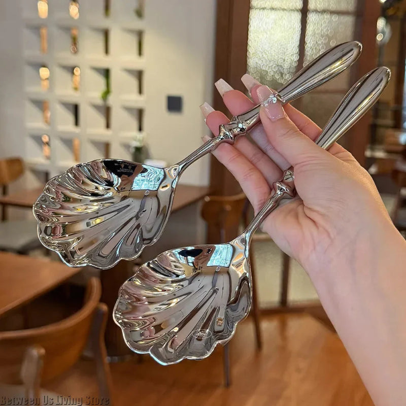 A hand holding a pair of ornate, polished silver serving spoons with scalloped, shell-shaped heads.