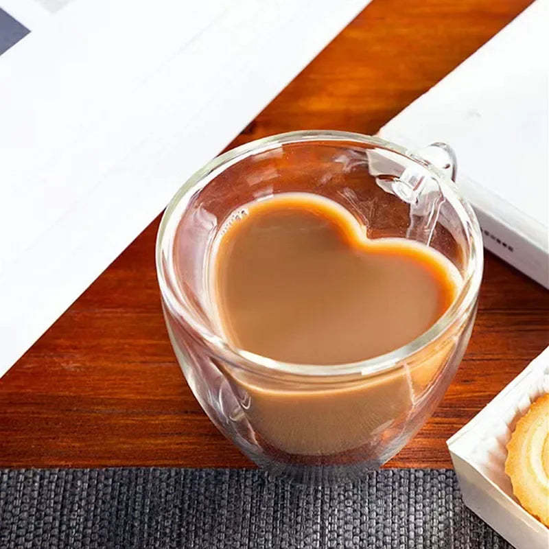 A glass, heart-shaped mug of coffee sits on a wooden table next to a book and a box of cookies.