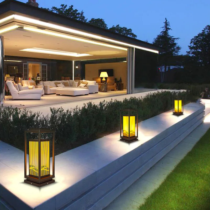 Three modern, dark-framed outdoor lanterns with vertical slats emit a warm yellow glow along a tiered walkway at dusk. The walkway leads to a modern house with large sliding glass doors revealing a brightly lit interior with white furniture. Lush green hedges line the walkway, and the sky is a deep twilight blue.