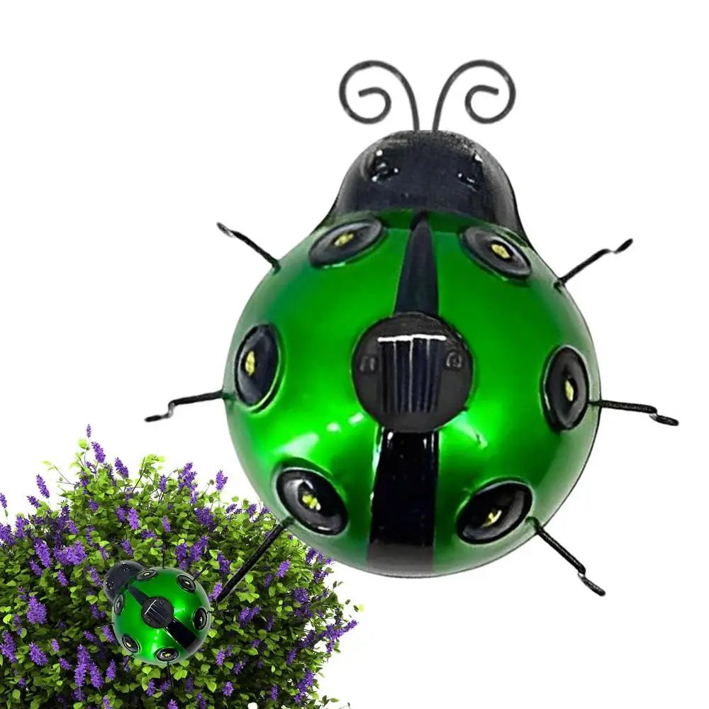A decorative green and black metal ladybug solar light is shown against a white background. It has black antennae and six black legs, with small lights embedded in its spots and a solar panel on its back. A smaller version of the ladybug is near a purple flowering plant.