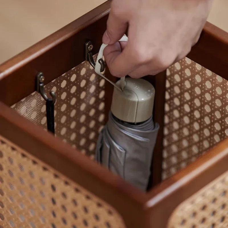 Close-up shot of a hand placing a folded, silver umbrella into a square, dark wood umbrella stand with light rattan weave sides. Metal hooks are visible inside the stand.