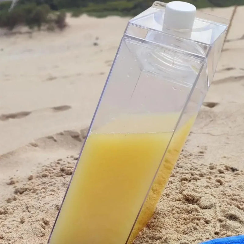 A clear, carton-shaped jug half-filled with pineapple juice, stuck in the sand at a beach.