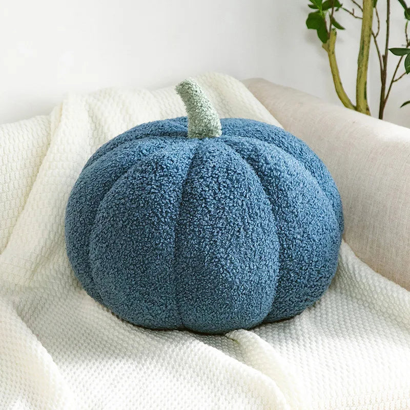 A dark blue, fuzzy, pumpkin-shaped pillow with a light green stem rests on a white textured blanket on a couch.