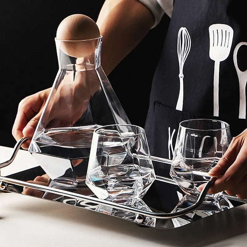 Hands holding a silver tray with a clear glass pitcher and two matching faceted glasses filled with water. The person is wearing a dark apron with kitchen utensil graphics.