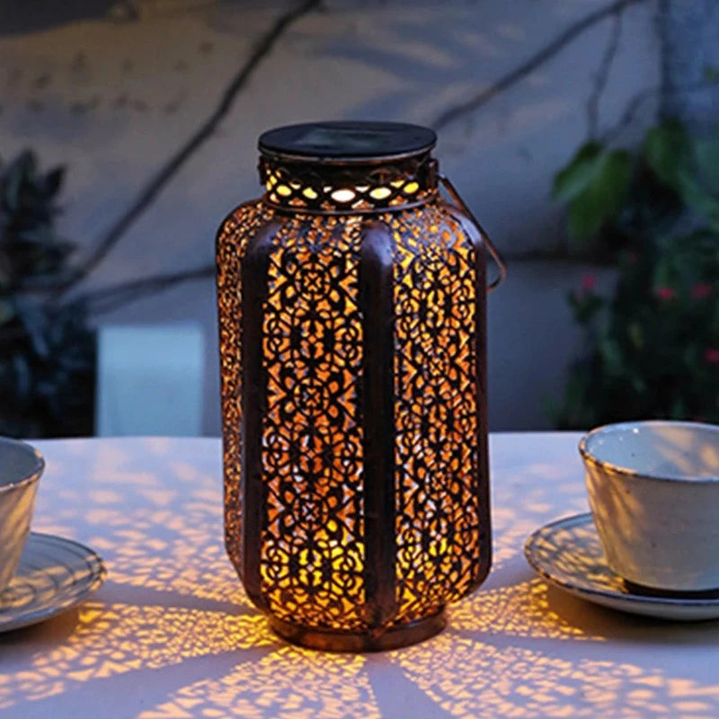 A decorative metal solar lantern with intricate cutouts emits a warm glow on a white tablecloth, casting patterned shadows. Two teacups and saucers are partially visible on either side. The background is softly blurred, showing outdoor elements.