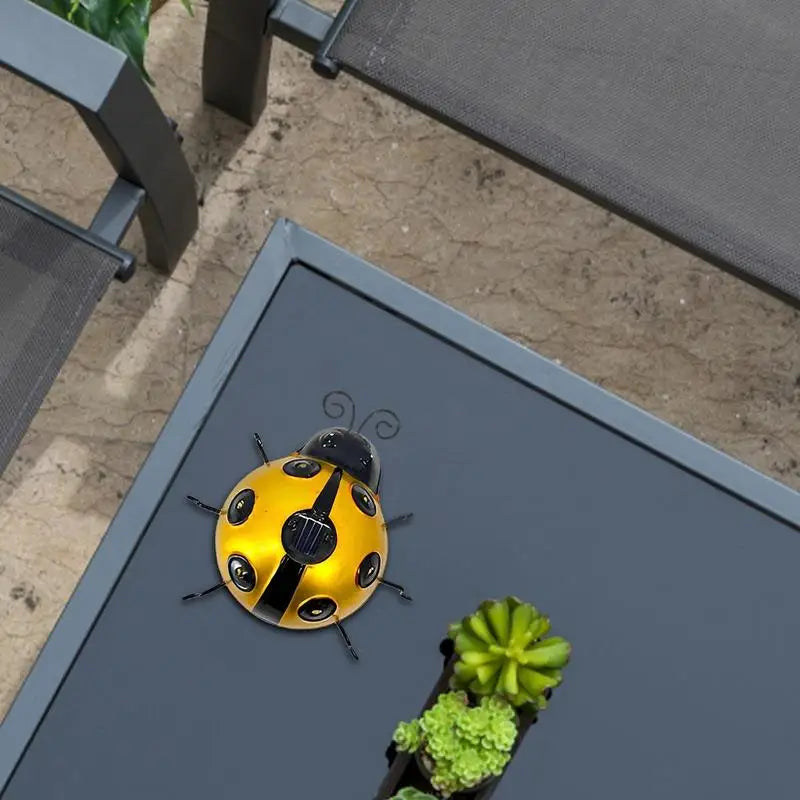 A decorative yellow metal ladybug with black stripes and antennae is attached to the edge of a gray outdoor table. A small potted succulent is also on the table. Parts of outdoor furniture are visible.