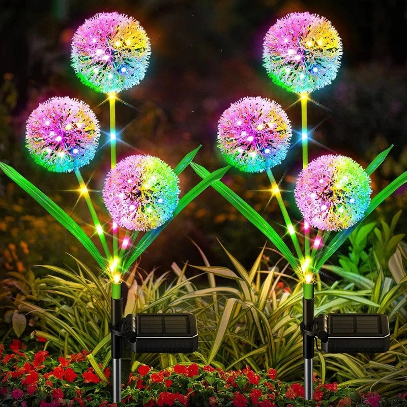 Six solar-powered garden lights are arranged in two rows. Each light features a spherical, dandelion-like head emitting a vibrant, multicolored glow, including pink, yellow, blue, and green. They have green, leaf-shaped accents and black solar panels at the base. The lights are placed among low-lying greenery and small red flowers.