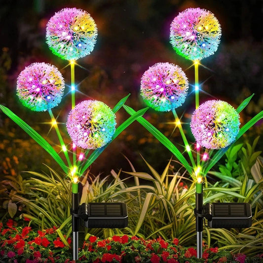 Six solar-powered garden lights are arranged in two rows. Each light features a spherical, dandelion-like head emitting a vibrant, multicolored glow, including pink, yellow, blue, and green. They have green, leaf-shaped accents and black solar panels at the base. The lights are placed among low-lying greenery and small red flowers.
