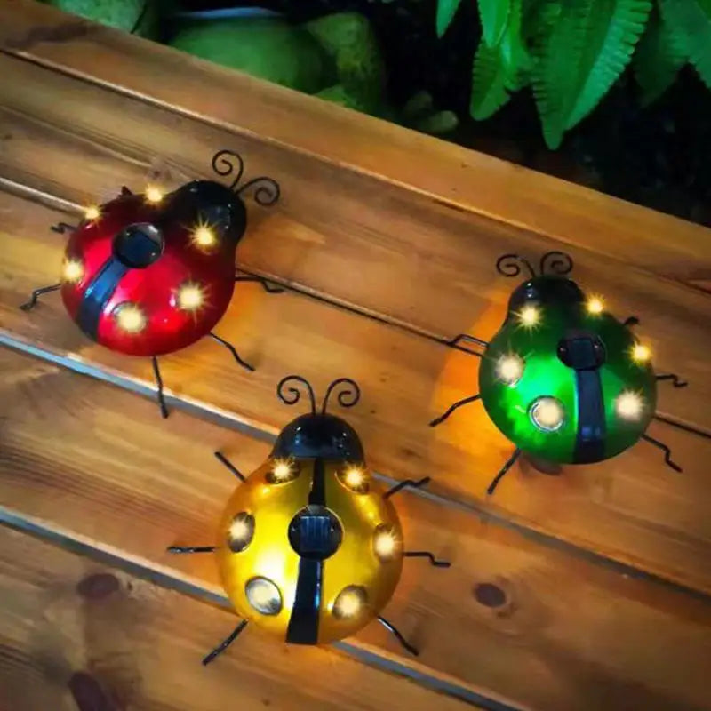 Three illuminated decorative ladybug solar lights, one red, one green, and one yellow with black stripes, sit on a wooden surface at night. Each ladybug has glowing spots and black antennae and legs.