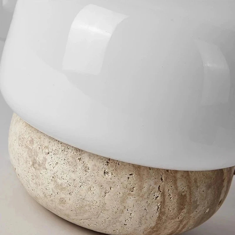 A close-up shot shows the base and lower portion of the shade of a small table lamp. The base is a textured, light-colored stone with visible pores and variations in tone. The shade is made of smooth, glossy white material that curves gently. A hint of the surface the lamp is resting on is visible at the bottom.