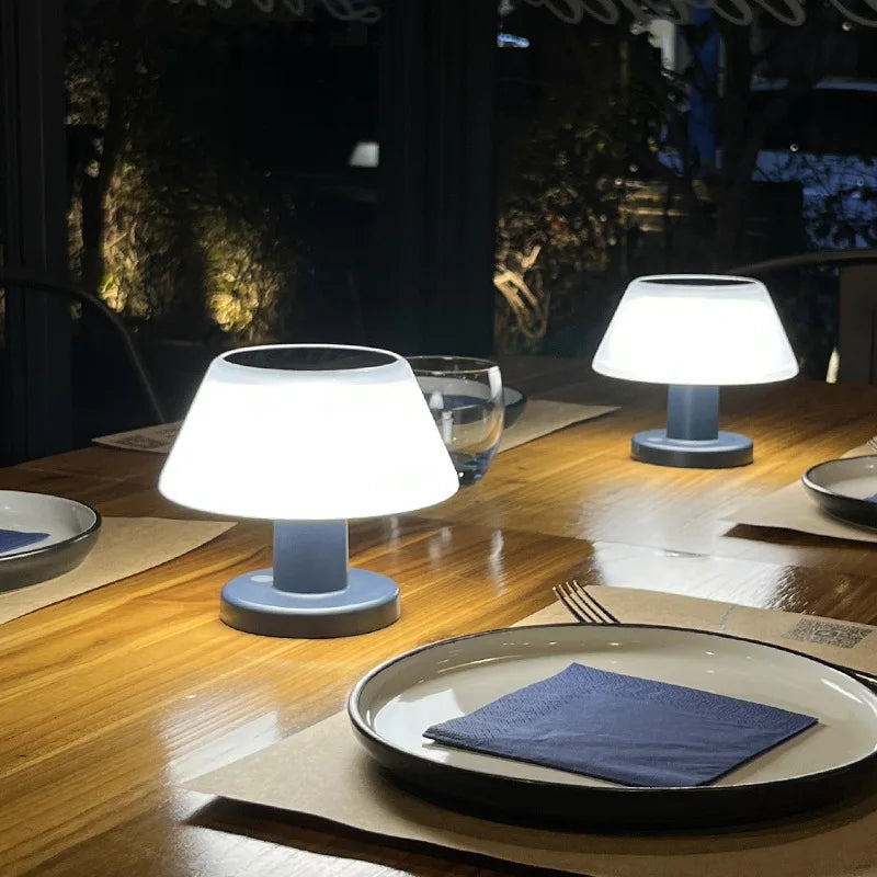 Two small, modern table lamps with white, slightly tapered shades and blue bases illuminate a wooden outdoor dining table set for two. Each place setting has a white plate with a blue napkin, a fork, and a drinking glass. The background is dark with blurred lights and greenery, suggesting a restaurant patio at night.
