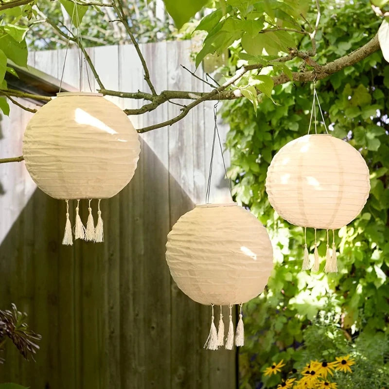 Three round, cream-colored solar lanterns with tassels hang from tree branches in a garden setting with green foliage and yellow flowers.