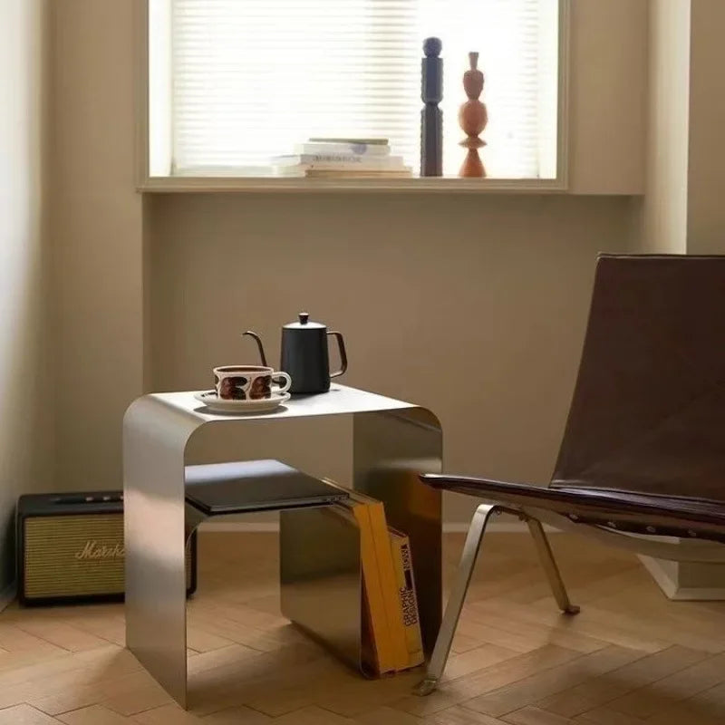 A metallic silver nightstand with rounded corners holds a white lamp, books, and other decorative items.