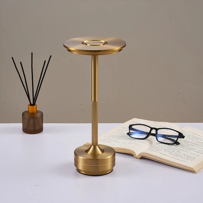 "A modern gold-colored desk lamp with a circular flat top and a textured handle, placed on a white table. An open book with black-framed glasses rests nearby, and a reed diffuser in a brown glass container is positioned in the background.