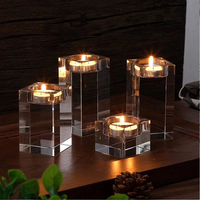Four clear, multifaceted crystal candle holders of varying heights, each containing a lit tea light candle, are arranged on a dark wooden surface. The candles cast a warm glow, and the crystal reflects the light. The background is a dark wooden wall. A pinecone is partially visible in the lower right corner.