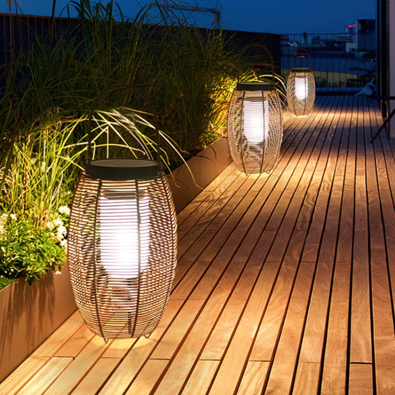 Three modern, oval outdoor lanterns made of dark metal slats emit a bright white glow on a wooden deck at dusk. The lanterns have dark, flat tops, likely solar panels. Tall grasses grow along the edge of the deck.