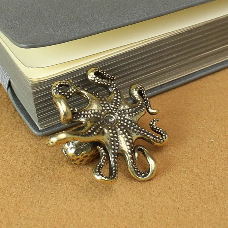 A small metallic octopus figurine with an antique brass or bronze finish, shown from the underside. The detailed design includes textured tentacles and a central circular embellishment. The figurine is placed near a closed gray notebook on a brown surface, giving it a vintage or steampunk aesthetic. It appears to be a decorative item, possibly a charm, ornament, or small accessory.