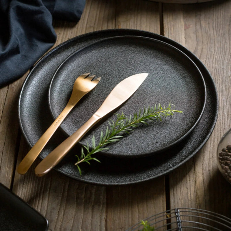 A stack of two round, matte black plates on a rustic wooden table, with a gold knife, fork, and a sprig of rosemary.