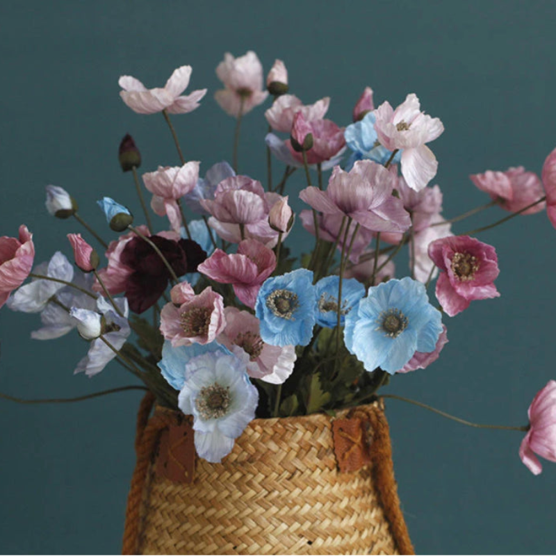 A wicker basket holds a bouquet of various colored poppy flowers, including shades of pink, purple, and blue, against a dark teal background.