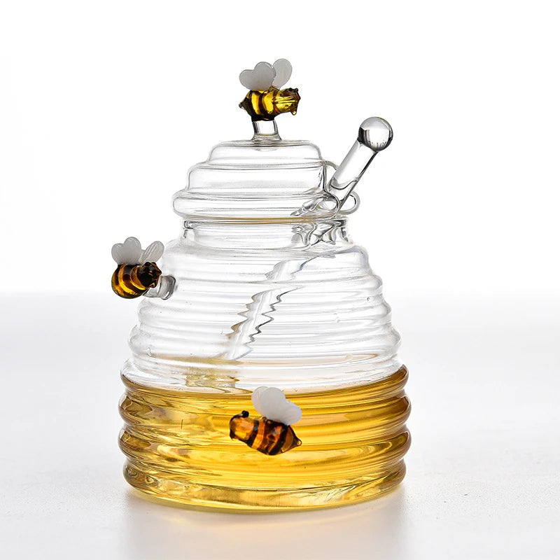 A clear glass honey jar shaped like a beehive with a lid, dipper, and three bee accents, partially filled with honey, on a white background.