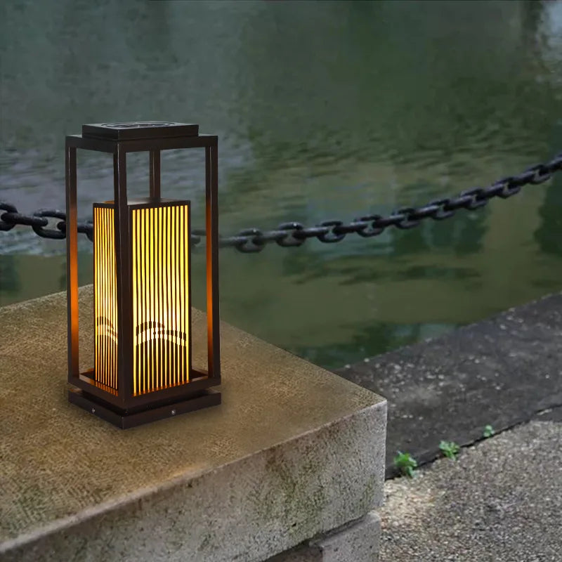 A modern, dark-framed outdoor lantern with vertical slats emits a warm yellow glow as it sits on a stone ledge beside a body of water with a chain railing.
