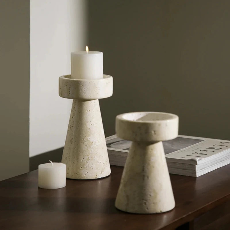 Two textured, light-colored faux travertine stone candle holders of different heights, one holding a lit white pillar candle, arranged on a dark wooden surface with a stack of magazines.