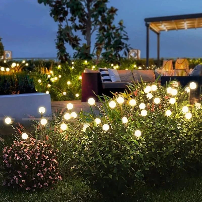 Numerous slender black stems with glowing warm white spherical lights top lush greenery in a garden at dusk. The lights are scattered throughout the foliage, creating a magical, firefly-like ambiance. In the blurred background, outdoor furniture with cushions and a pergola are visible under the evening sky.