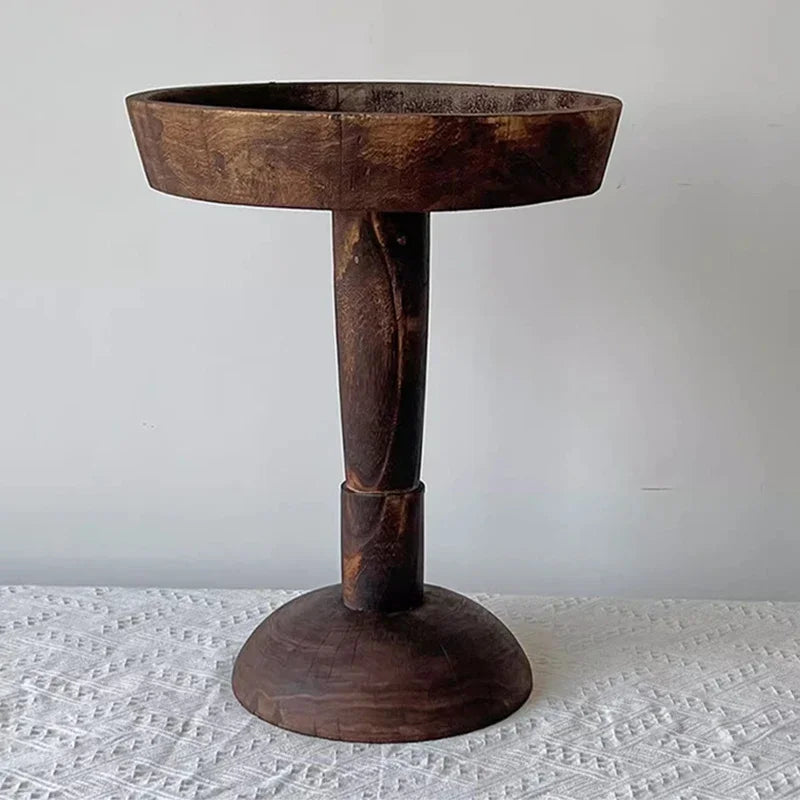 A rustic, dark wood, round side table (Momo Wabi-sabi Log Small Coffee Table) with a shallow tray top, standing on a patterned tablecloth against a neutral background.