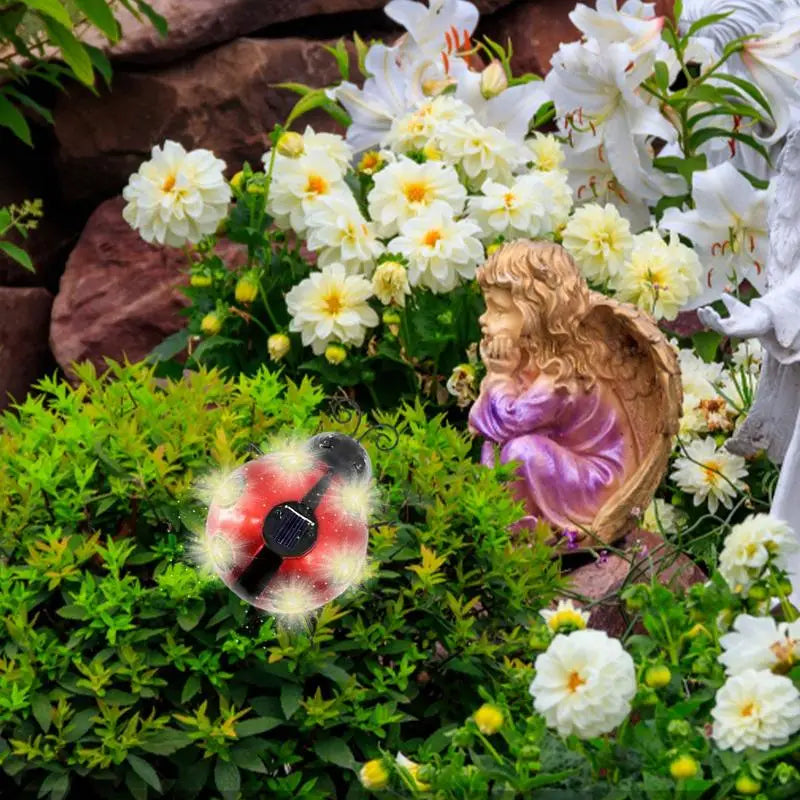 A decorative solar-powered ladybug spinner with red, white, and black elements is placed among green plants and white flowers in a garden setting. A small angel statue with a purple robe and golden wings is nearby.